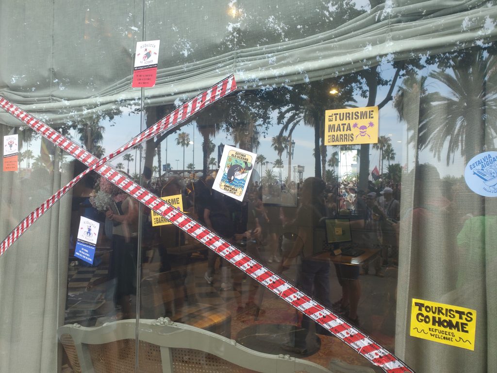 Aufschriften der Sticker: „Tourist go home, refugees welcome“, „Tourismus tötet Nachbarschaften“ und „Liebe die Ozeane, versenke die Reichen“ – der Sticker zeigt einen Wal, der ein Kreuzfahrtschiff versenkt.