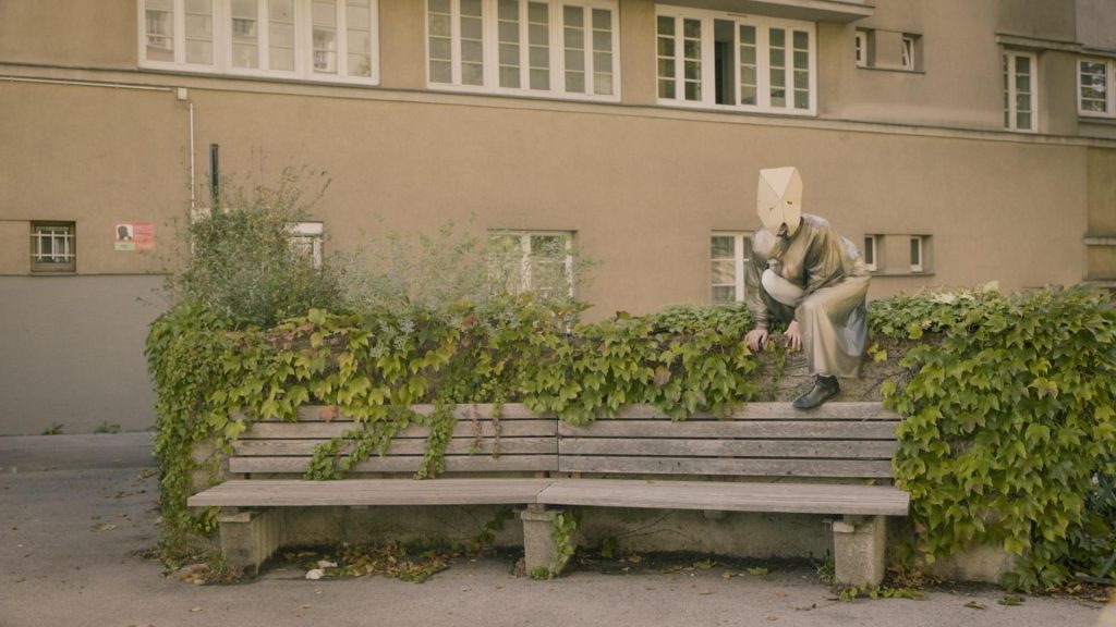 Ein*e Schauspieler*in mit Pappmaske hockt auf einer Mauer vor einer Bank