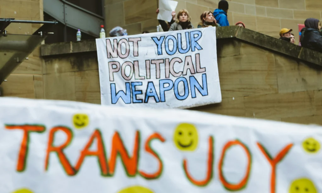 Eine Gruppe von Menschen mit einem Banner, auf dem in den Farben der Trans Flagge steht "not your political weapon". Davor ein anderes Banner mit der Aufschrift "Trans Joy" und Smileys.